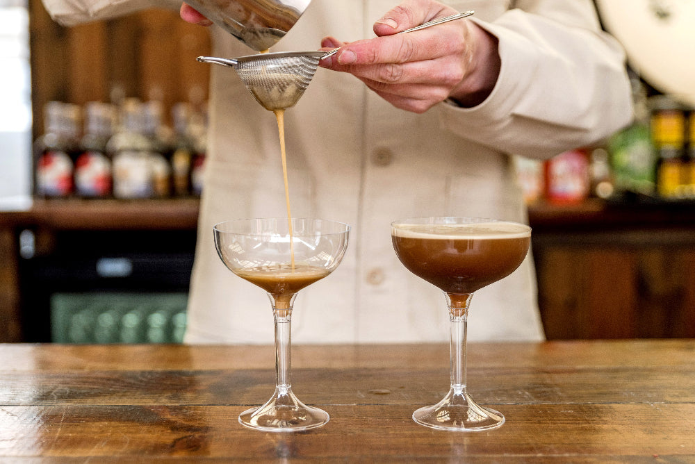Barista pouring a drink into two glasses on a wooden bar.