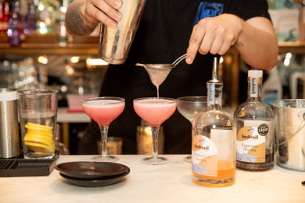 Bartender pouring a pink cocktail into a glass with a blurred bar background